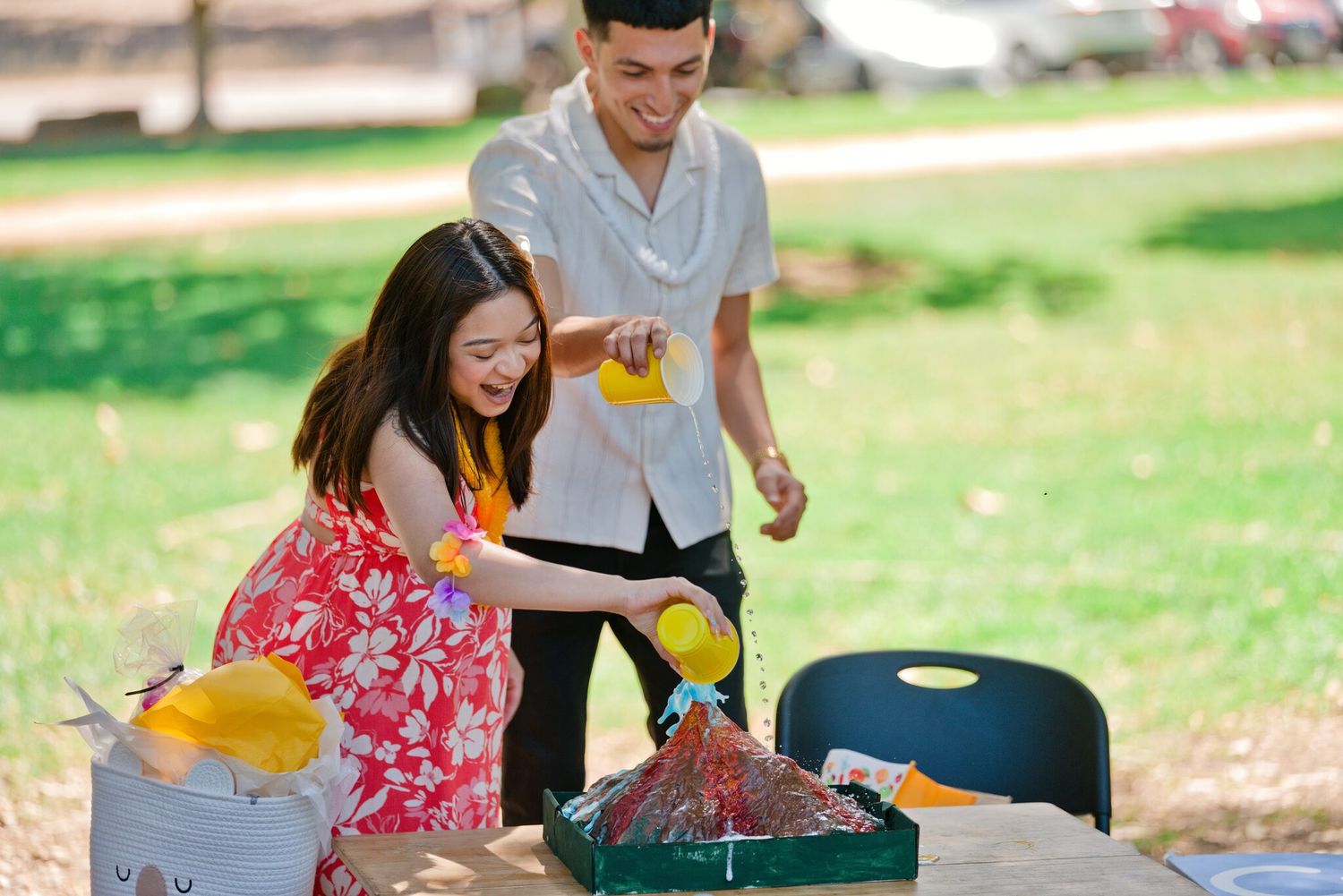 A smiling couple pours yellow liquid onto a colorful outdoor science project at a park table.