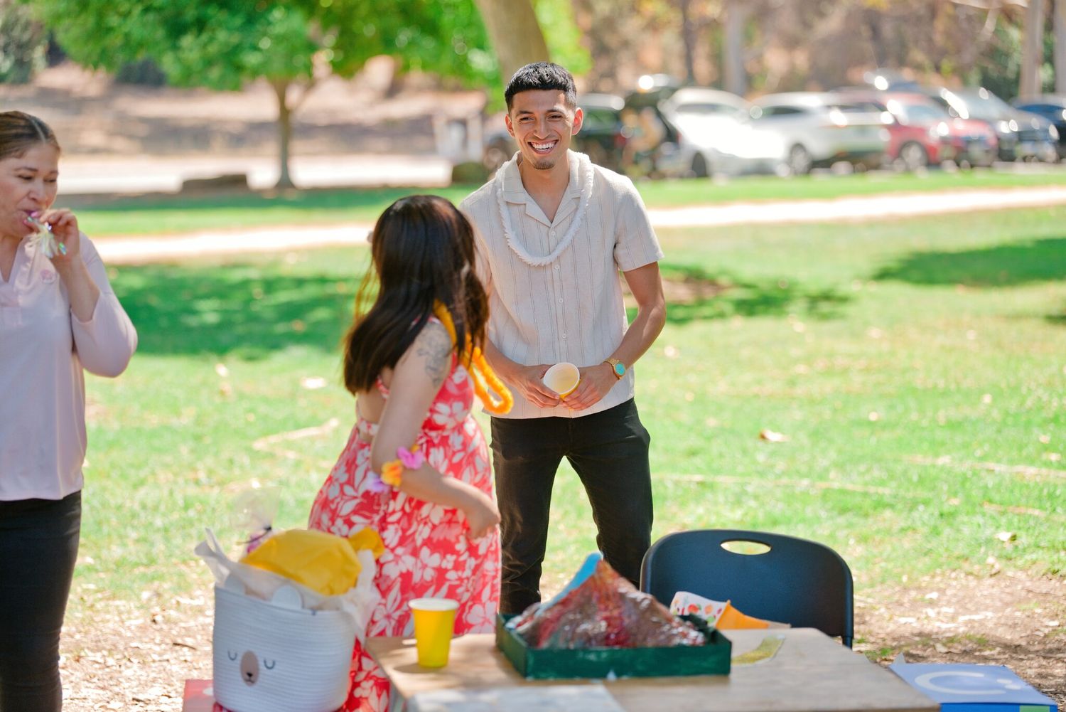 Young man and woman interact at picnic table during outdoor park celebration with food and drinks.