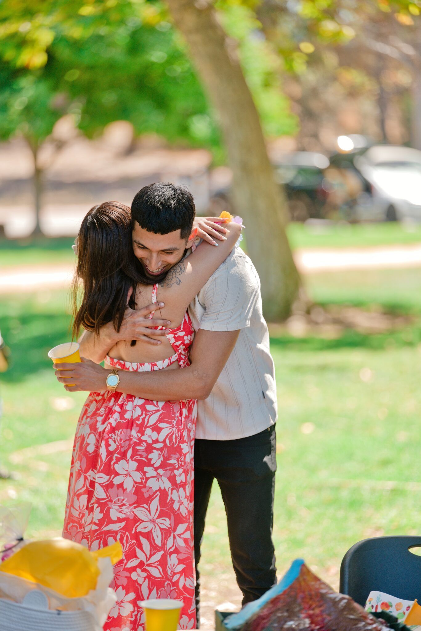 Couple hugging warmly at an outdoor park picnic on a sunny day, woman in red floral dress.