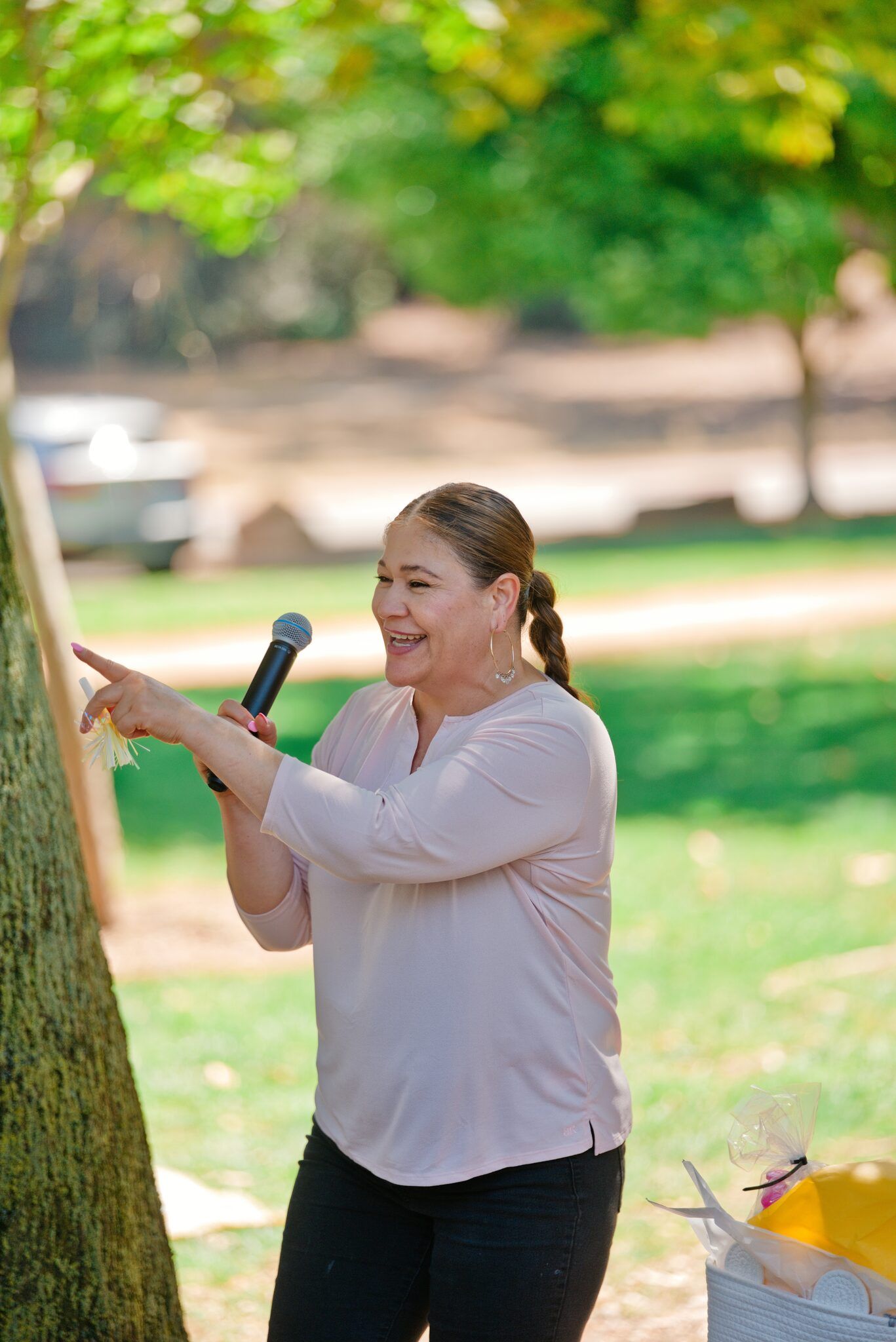 A cheerful woman holding a microphone points while speaking at an outdoor park gathering.
