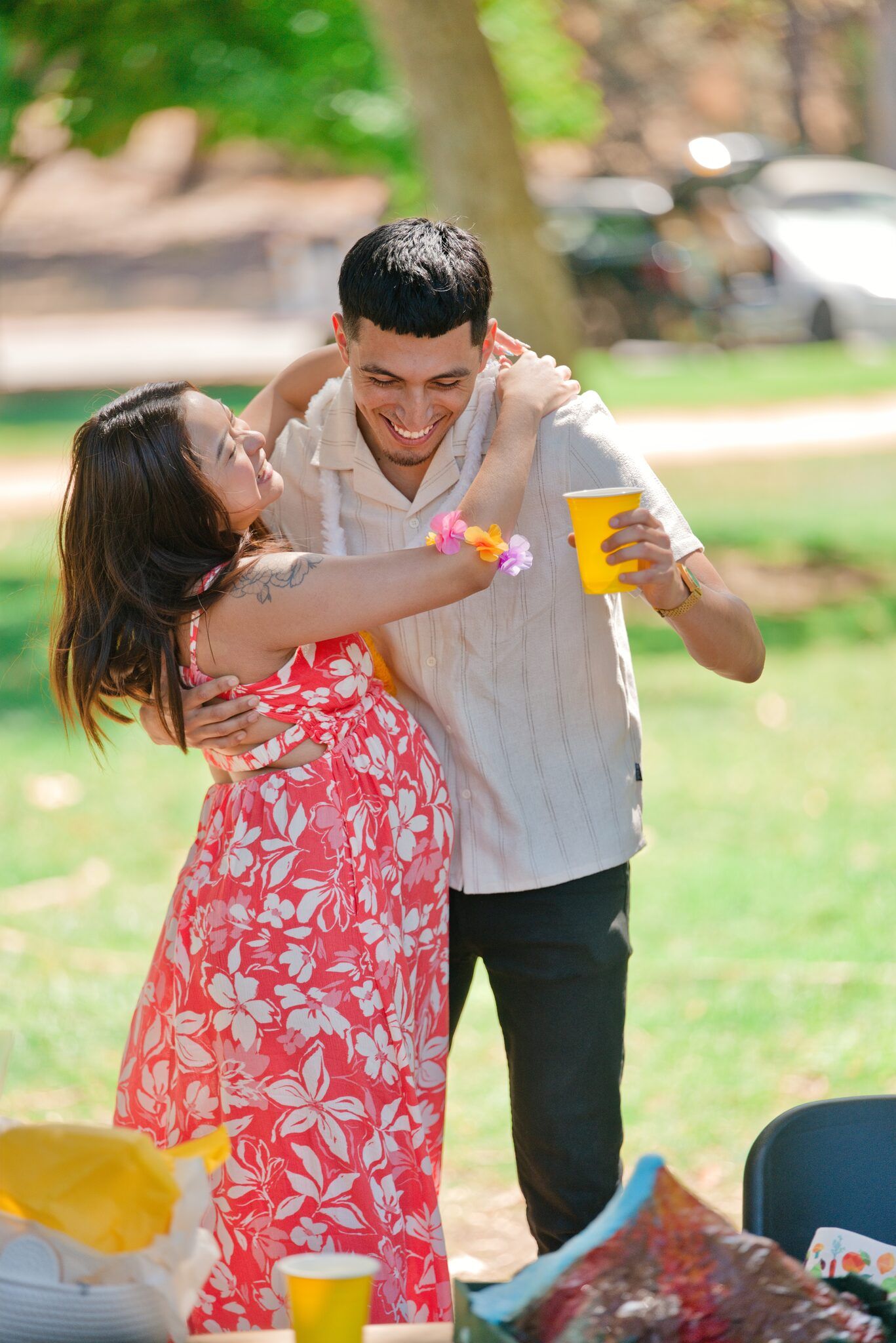 A smiling couple embraces at an outdoor park celebration with yellow cups and festive decor.