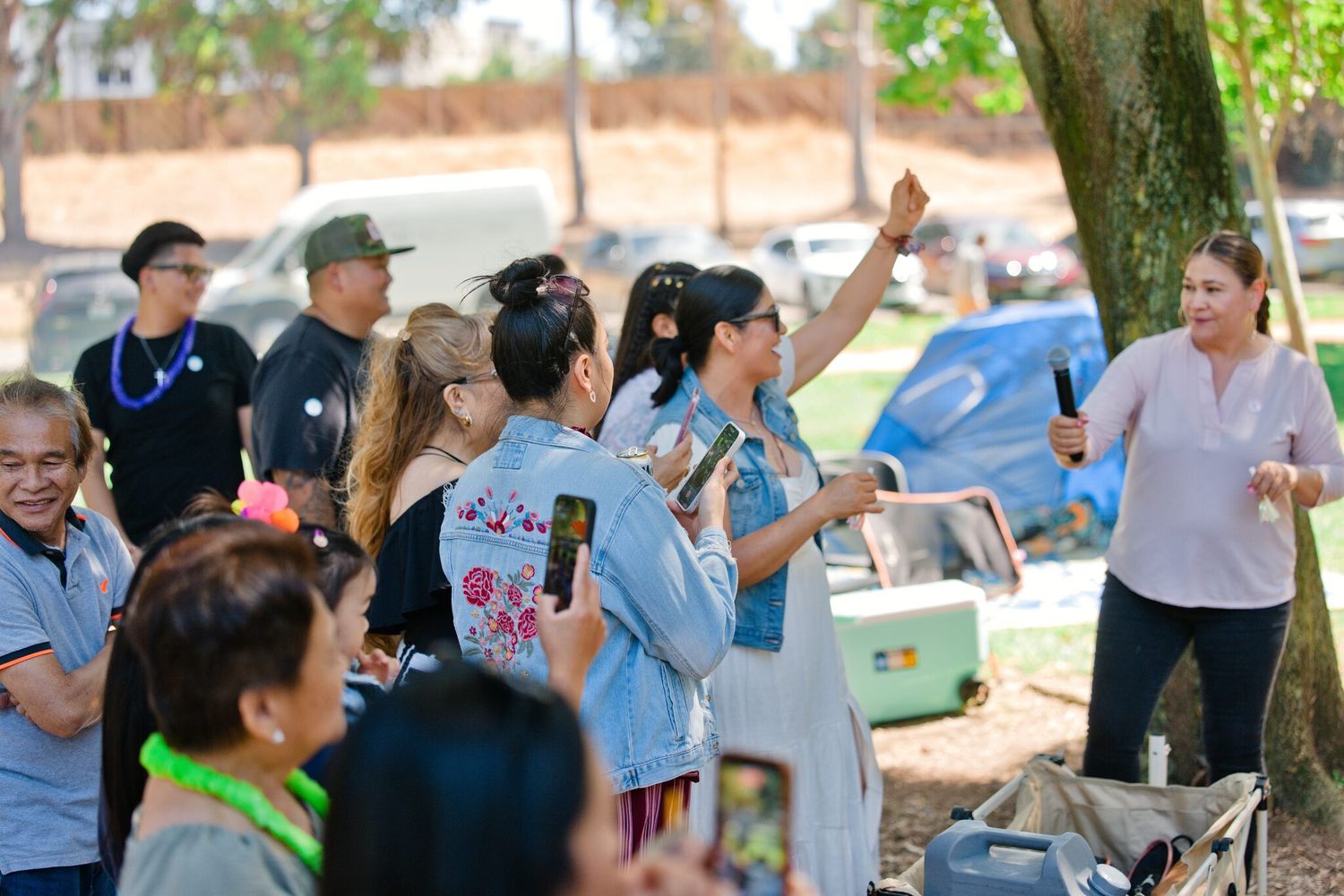 Group of people enjoying an outdoor gathering under trees, woman raising hand holding microphone.