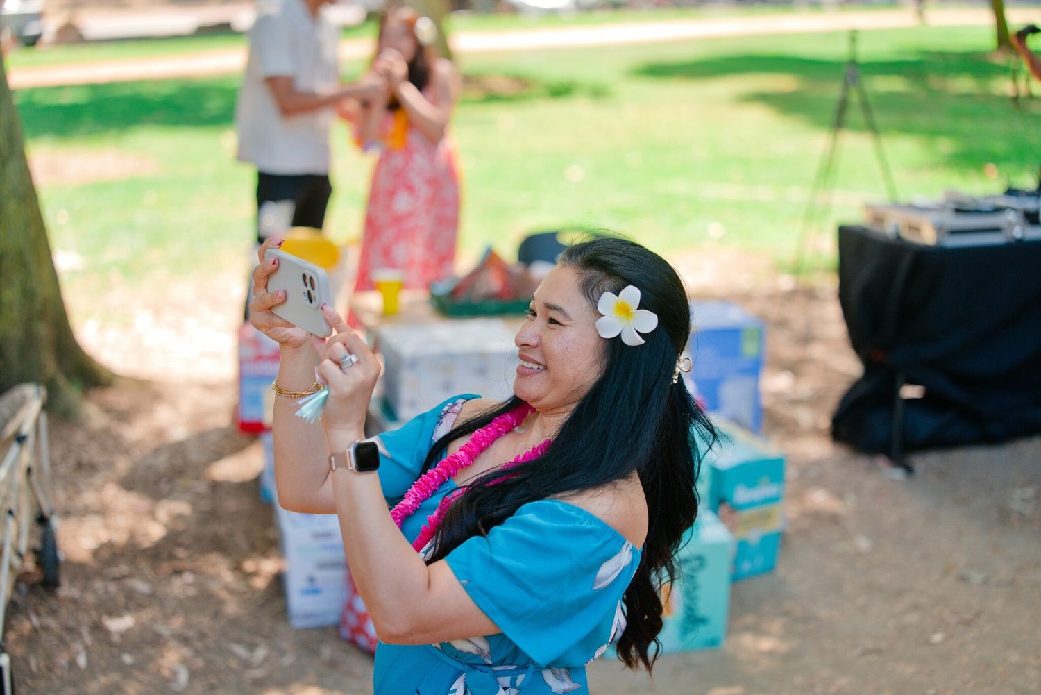 Woman with white flower in hair smiles while taking selfie at sunny outdoor park event.