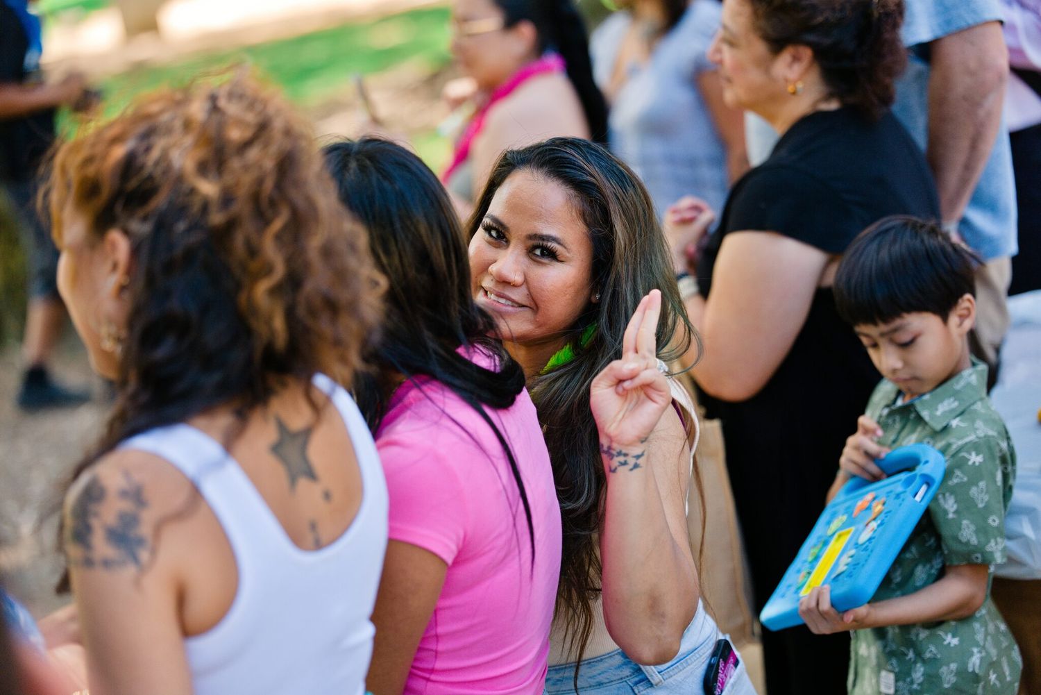 A woman in a pink shirt flashes a peace sign at a lively outdoor community park event.