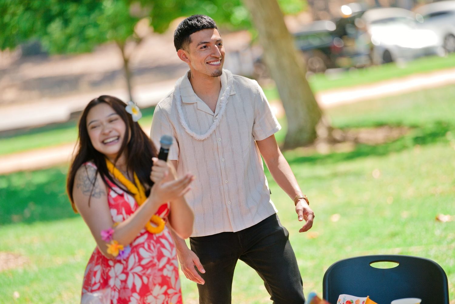 A laughing couple shares a microphone together at a sunny outdoor park celebration.