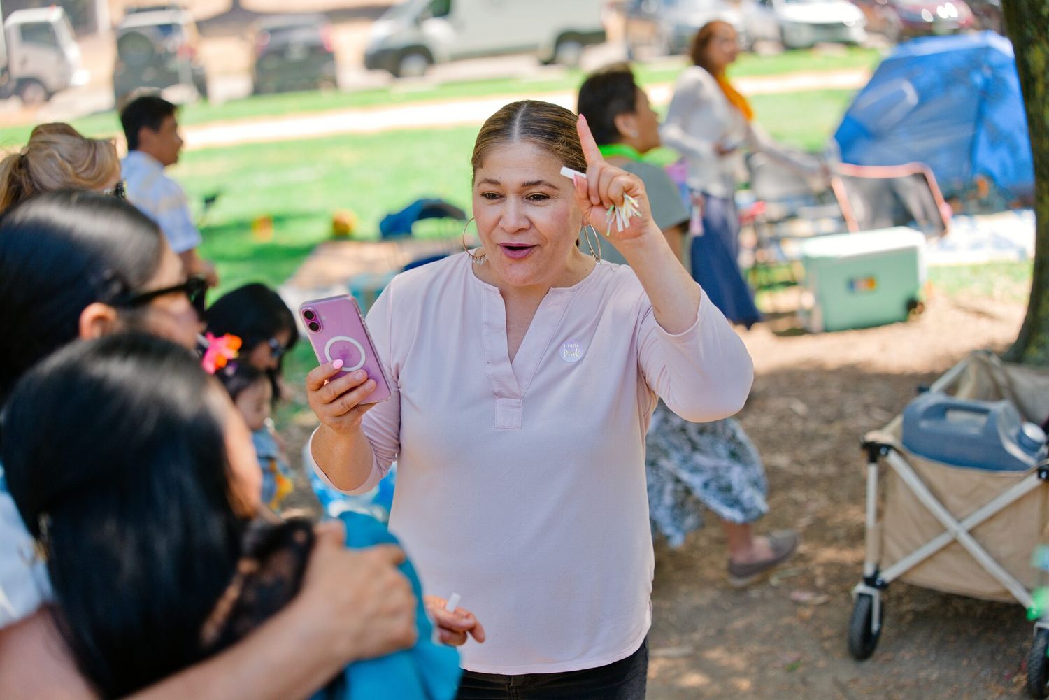 Woman holding a pink smartphone gestures expressively while socializing at an outdoor park event.