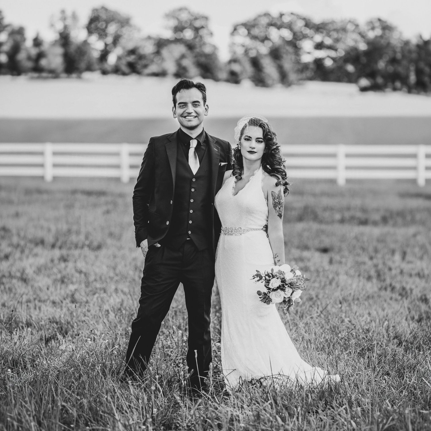 Black and white photo of a bride and groom posing in a grassy field with a white fence backdrop.