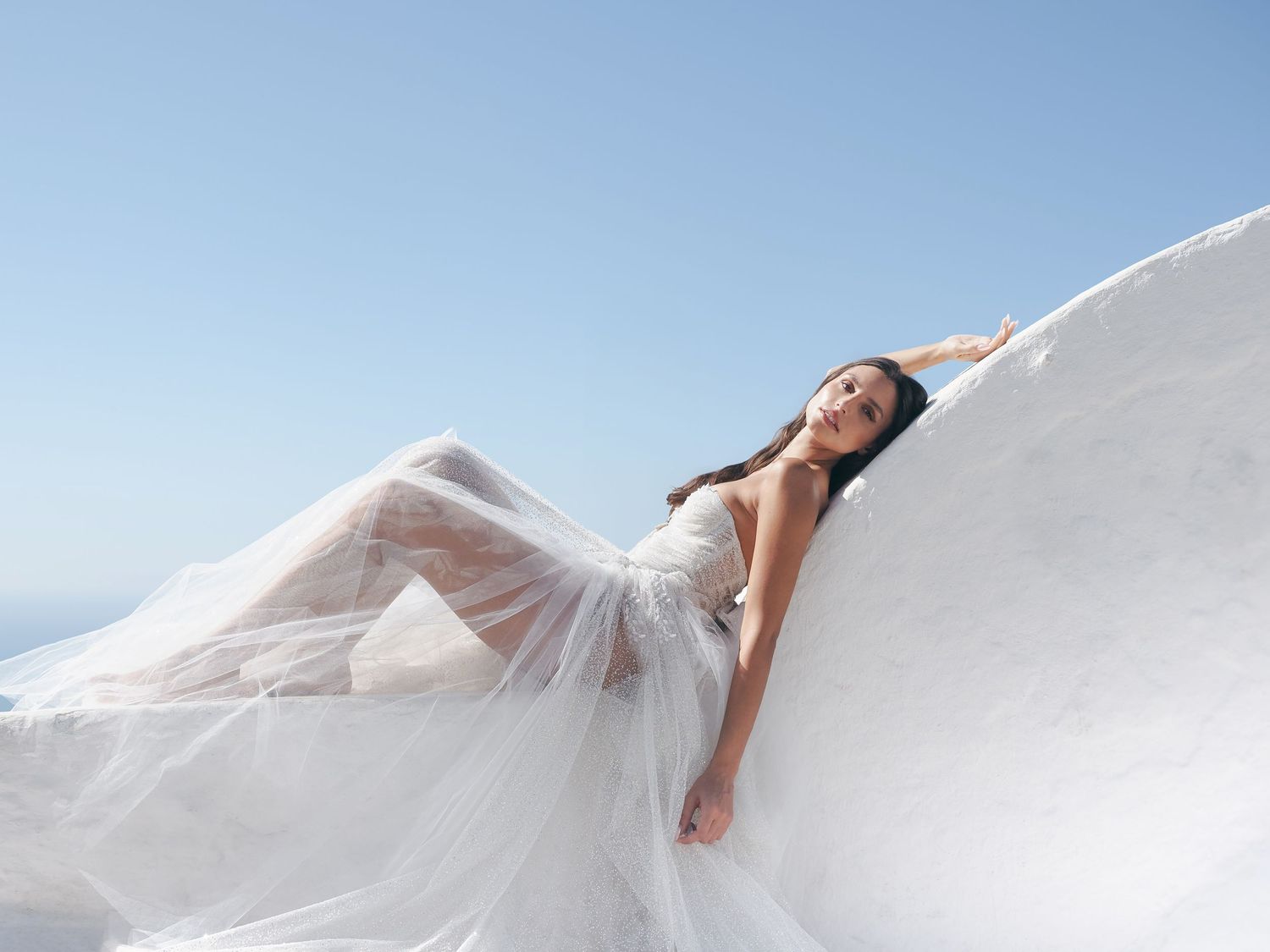 Woman in flowing white tulle gown poses gracefully against a serene blue sky backdrop.