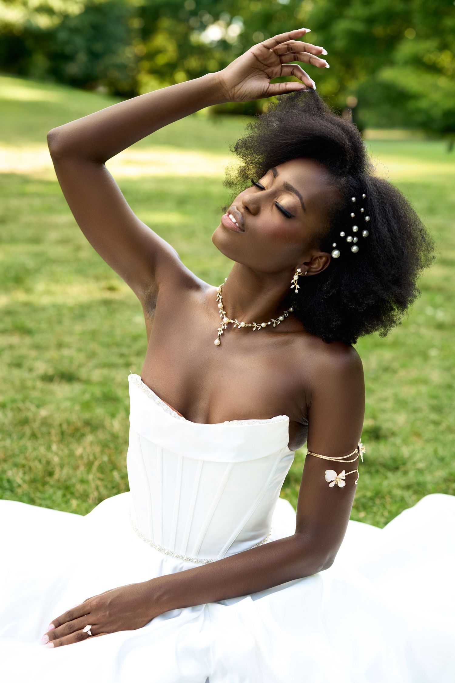 A Black bride in a white strapless gown sits gracefully outdoors, adorned with pearl jewelry and hair accessories.