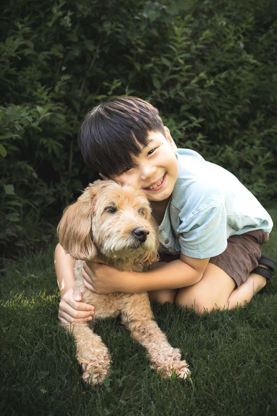 Smiling boy hugging a golden doodle dog on green grass with lush bushes in the background.