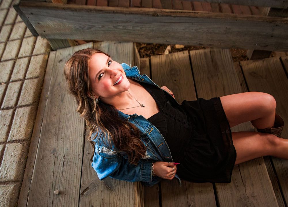 Young woman in denim jacket and black dress smiling on wooden deck steps in a professional portrait photo.