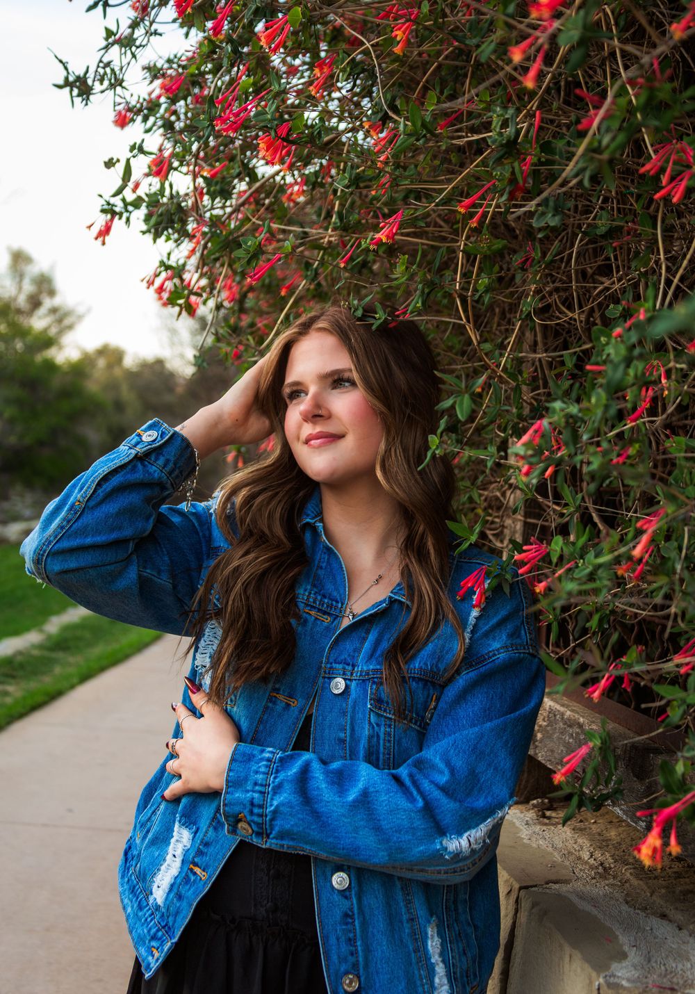 Young woman in distressed denim jacket poses by red flowering wall on a garden path.
