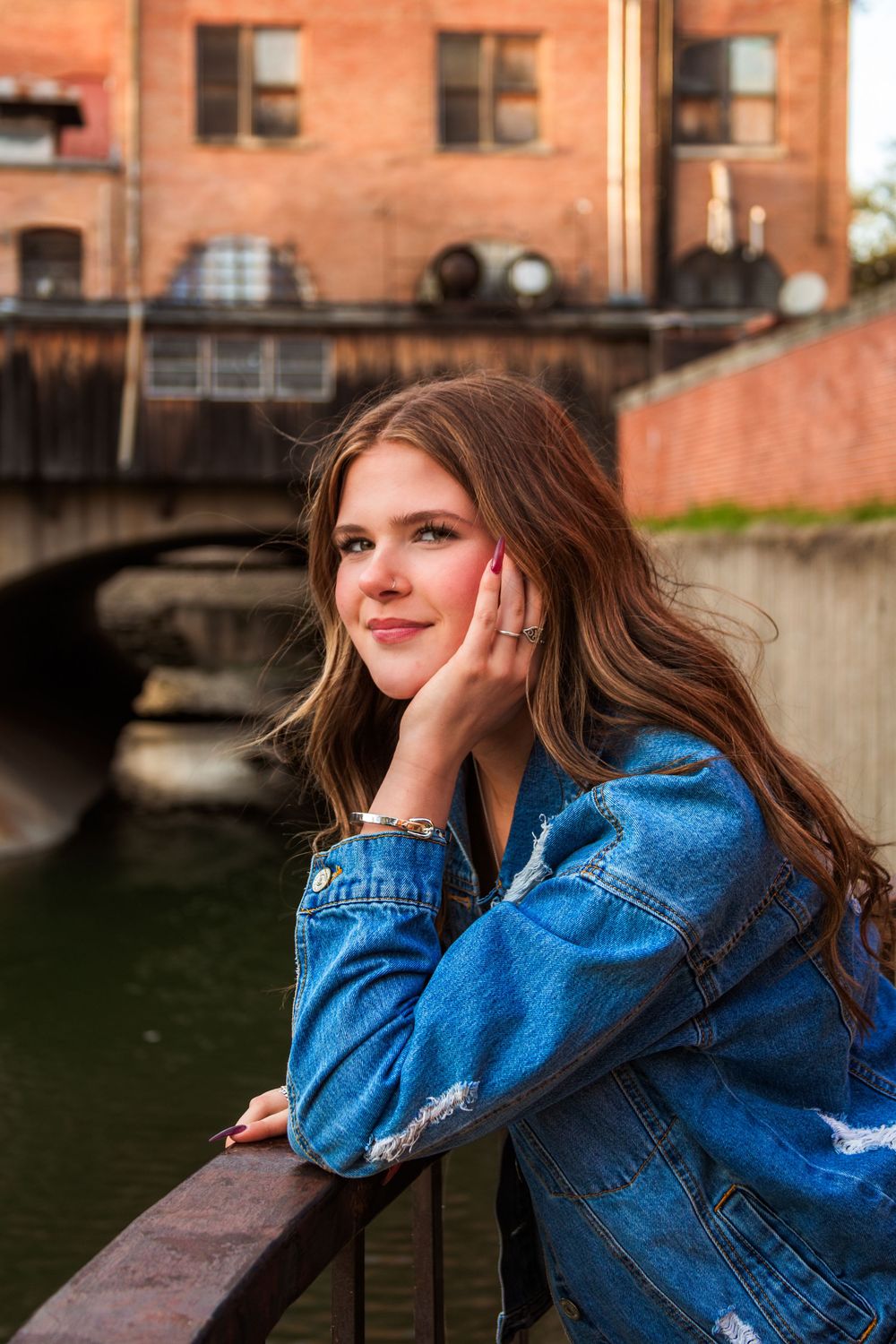 Young woman in denim jacket leaning on railing by urban canal, smiling softly with hand on cheek.