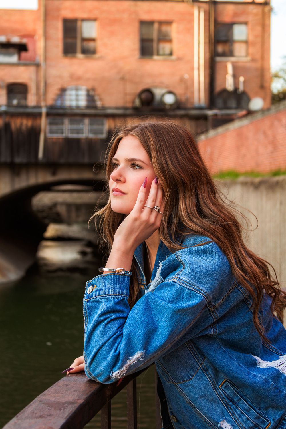 Young woman in denim jacket leaning on railing by urban canal with brick building and bridge in background.