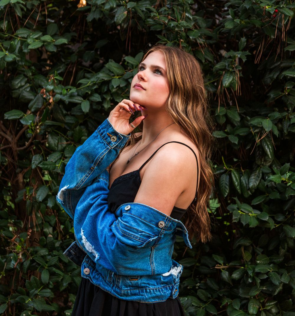 Young woman in denim jacket and black dress posing against lush green foliage backdrop.