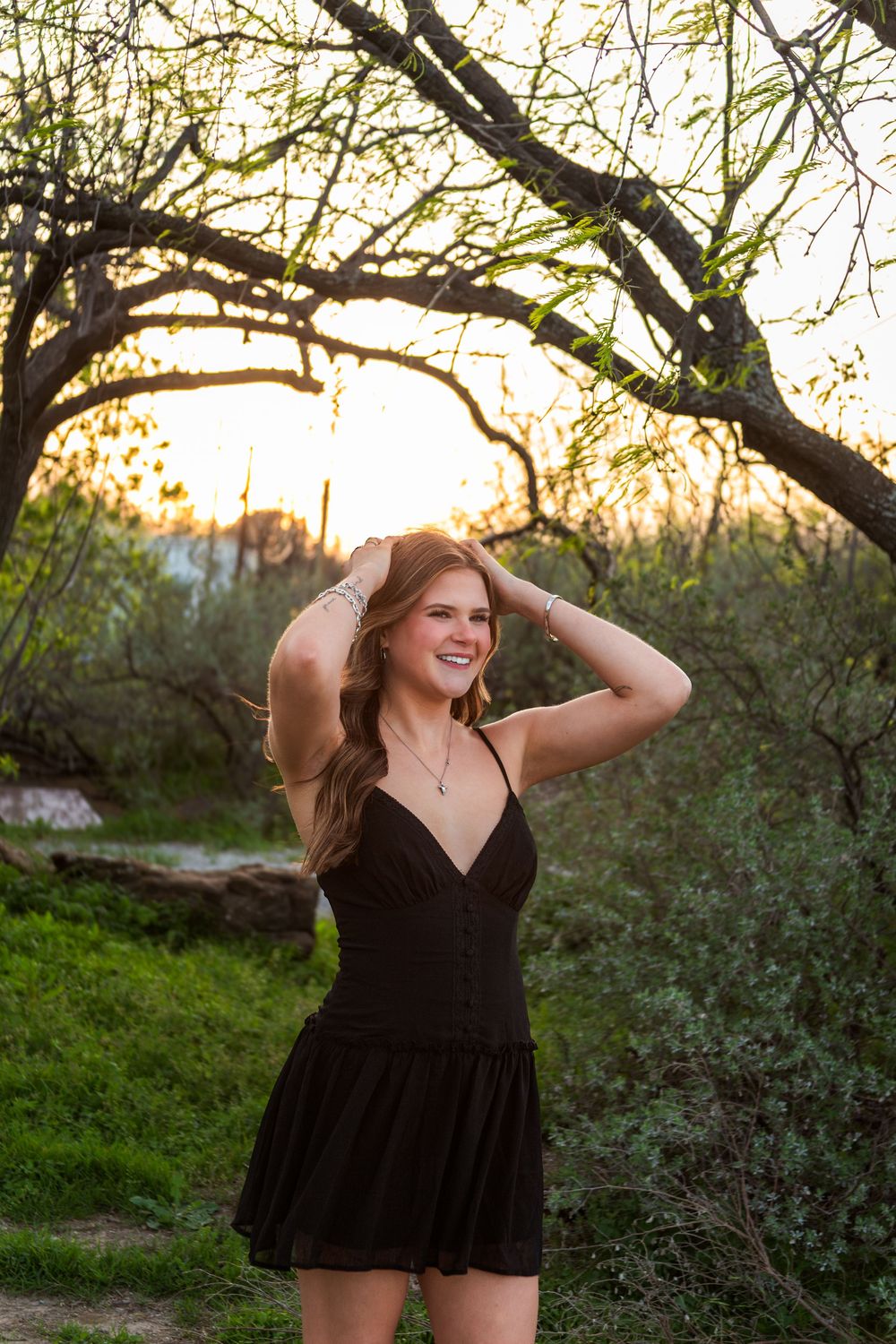 Young woman in black romper smiling outdoors at sunset, hands in hair, surrounded by trees and greenery.
