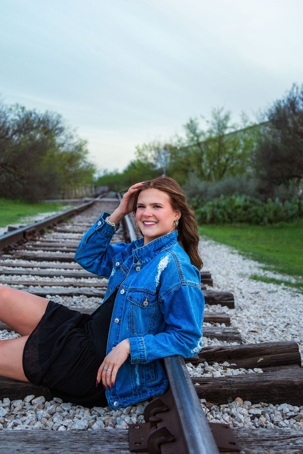 Young woman in denim jacket and black shorts smiling while sitting on railroad tracks surrounded by greenery.
