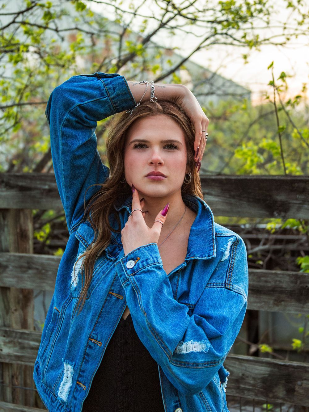 Young woman in distressed denim jacket poses outdoors near wooden fence with spring foliage in soft evening light.