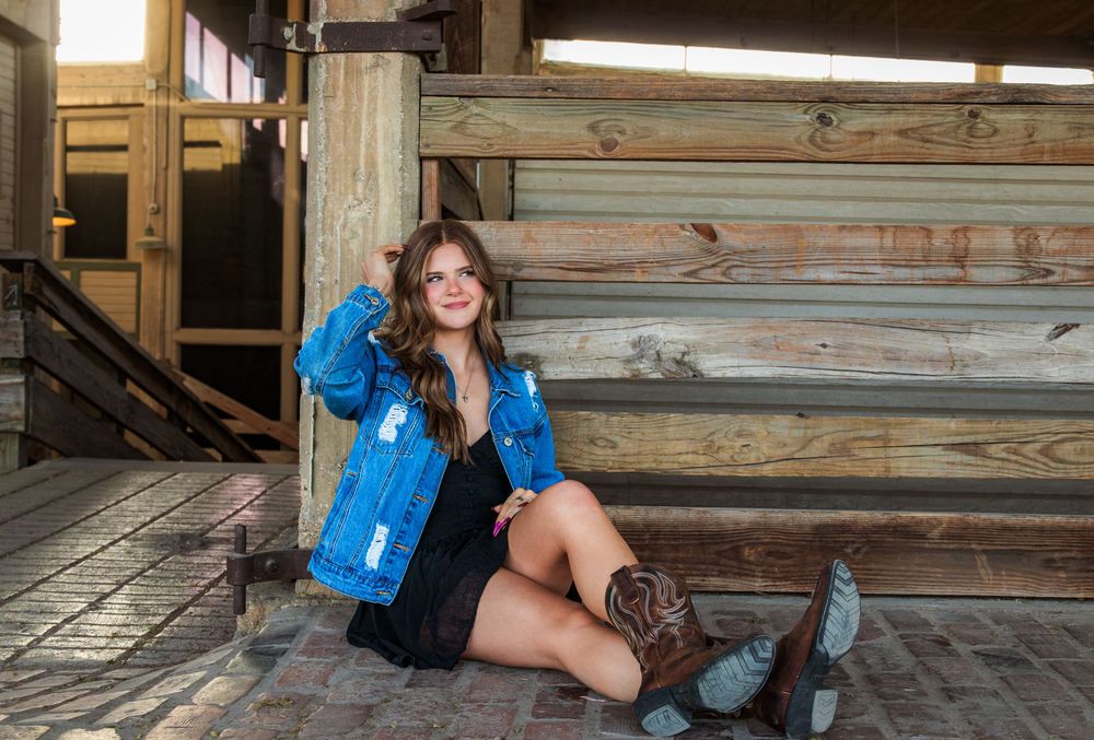 Young woman in denim jacket, black dress, and cowboy boots sits beside rustic log cabin porch.