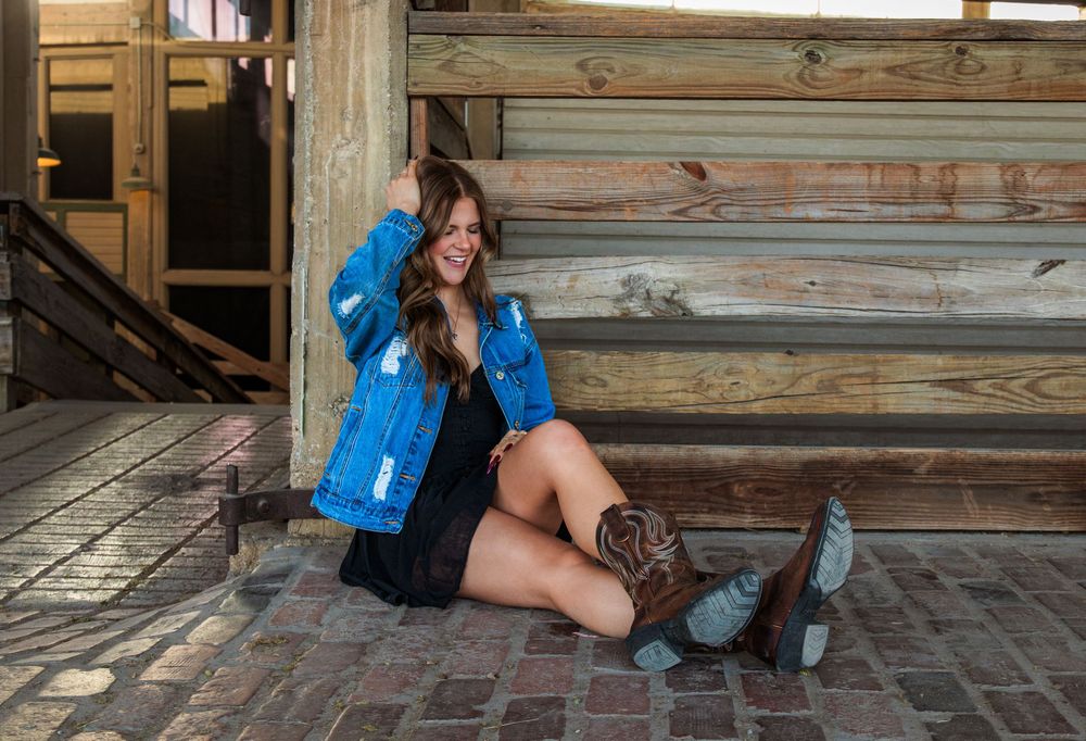 Young woman in denim jacket, black dress, and cowboy boots sitting on brick porch smiling.