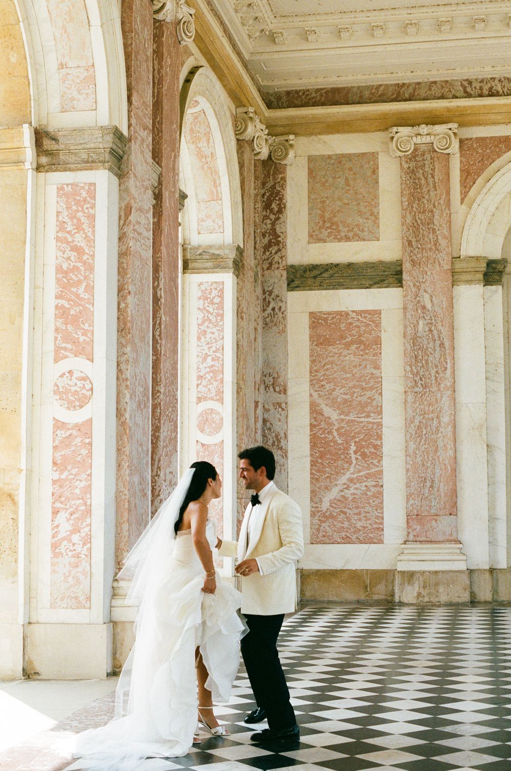 Bride and groom share a romantic moment in an elegant marble hall with pink columns and checkered floors. Best versailles photographer