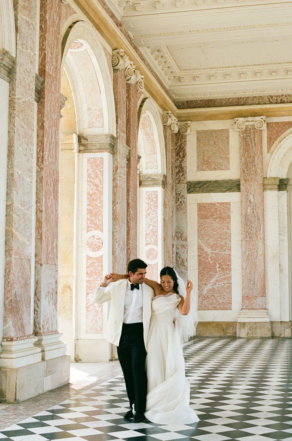 Bride and groom dancing in elegant marble hall with checkered floors and pink arched columns at Palace of Versailles.