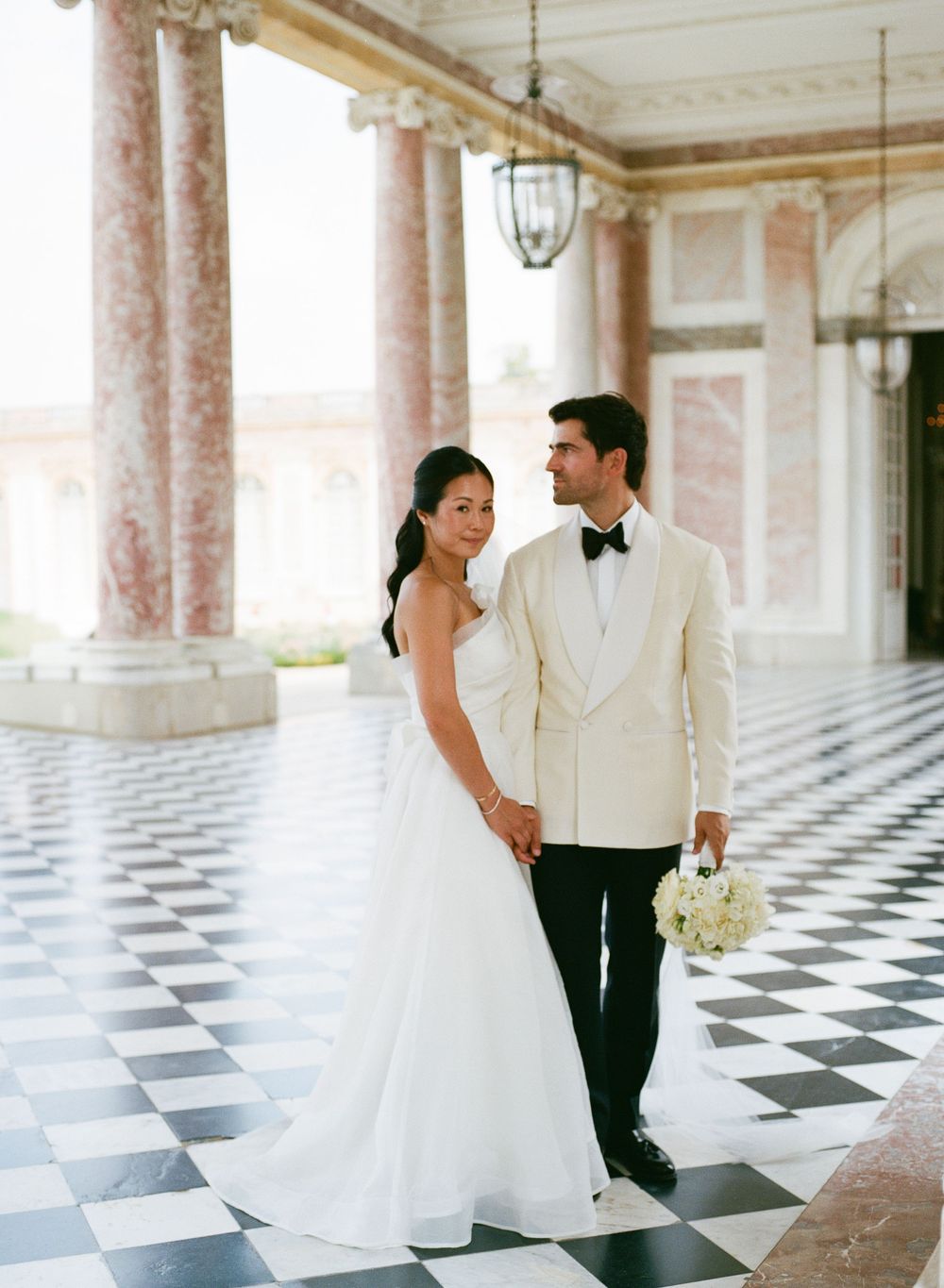 Elegant bride and groom pose on checkered marble floor with grand columns at Le Grand Trianon. Versailles wedding photographer, film photos