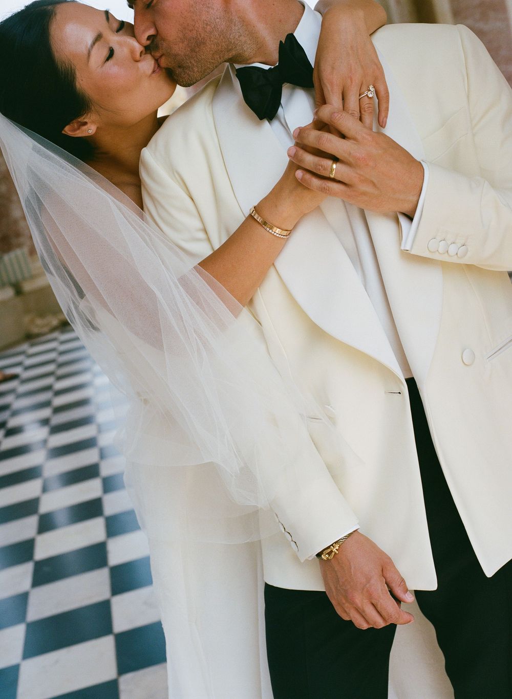 Bride in white veil and groom in ivory tuxedo share a romantic kiss at their Versailles wedding ceremony. Checkered floor at Le Grand Triano