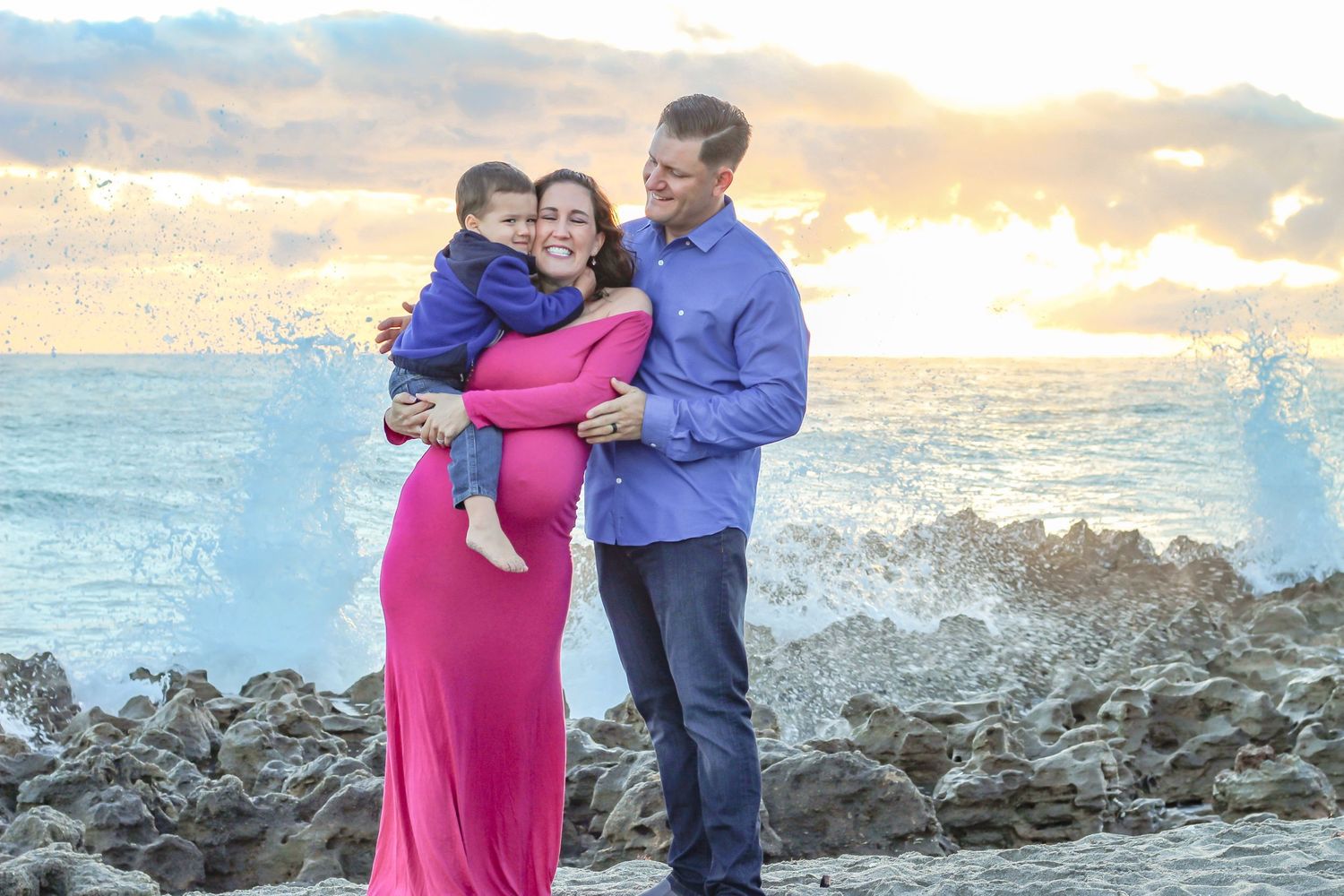Parents and toddler hugging during a maternity portrait session at Blowing Rocks Preserve in Jupiter, Florida as ocean waves crash behind.