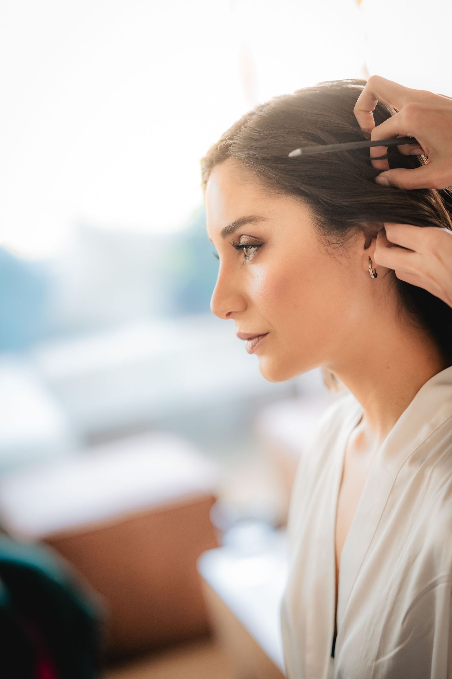 A bride in a white robe gets her dark hair styled by a hairdresser before her wedding ceremony.