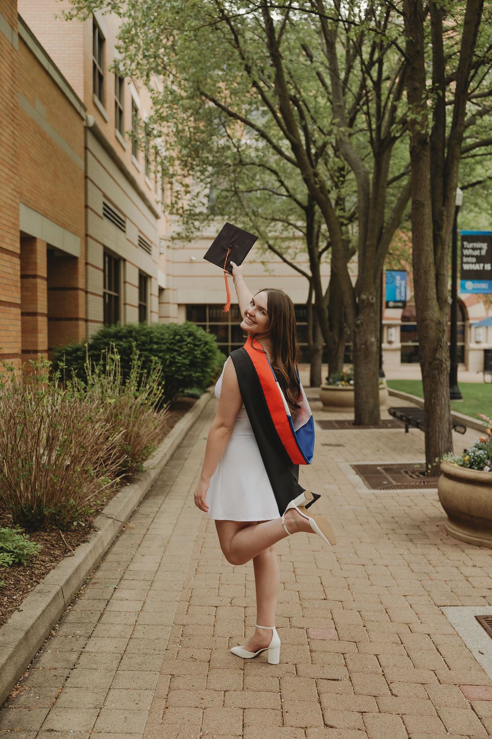 A joyful graduate in white dress and academic regalia holds cap aloft on a brick campus walkway.