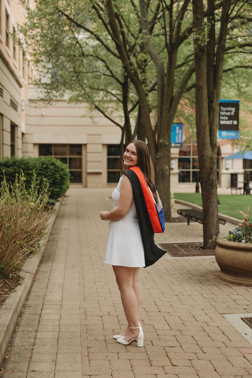 Smiling graduate in white dress and black cap gown poses on brick walkway surrounded by trees on college campus.
