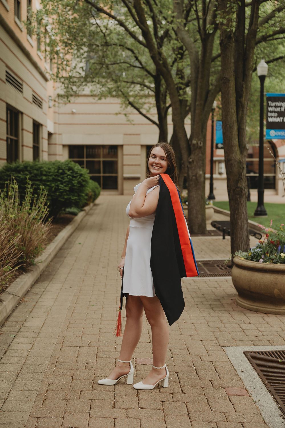 A smiling young woman in a white dress holds her black and orange graduation gown on a brick campus walkway.