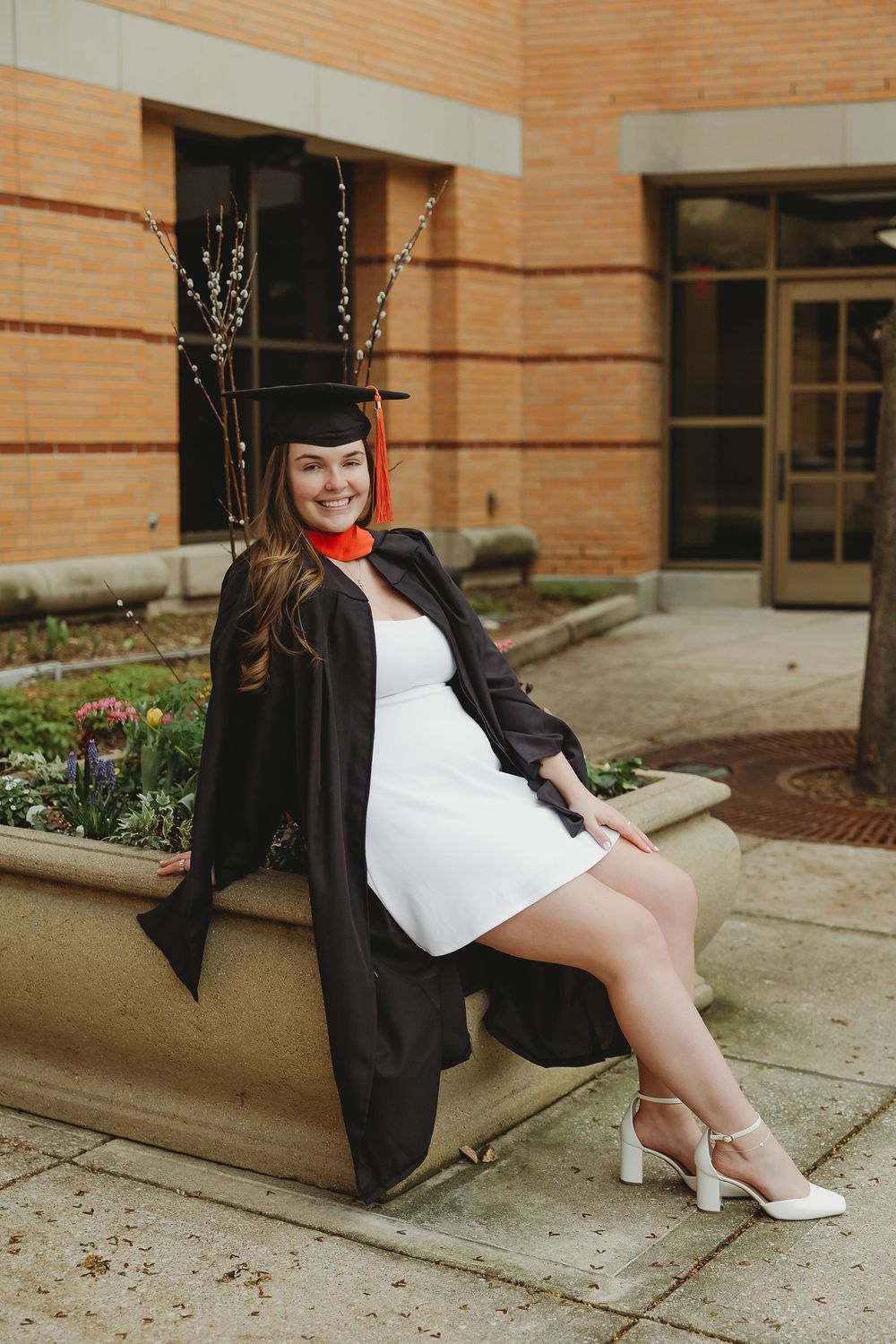 A smiling woman in black graduation cap and gown over white dress sits on stone ledge outside brick building.