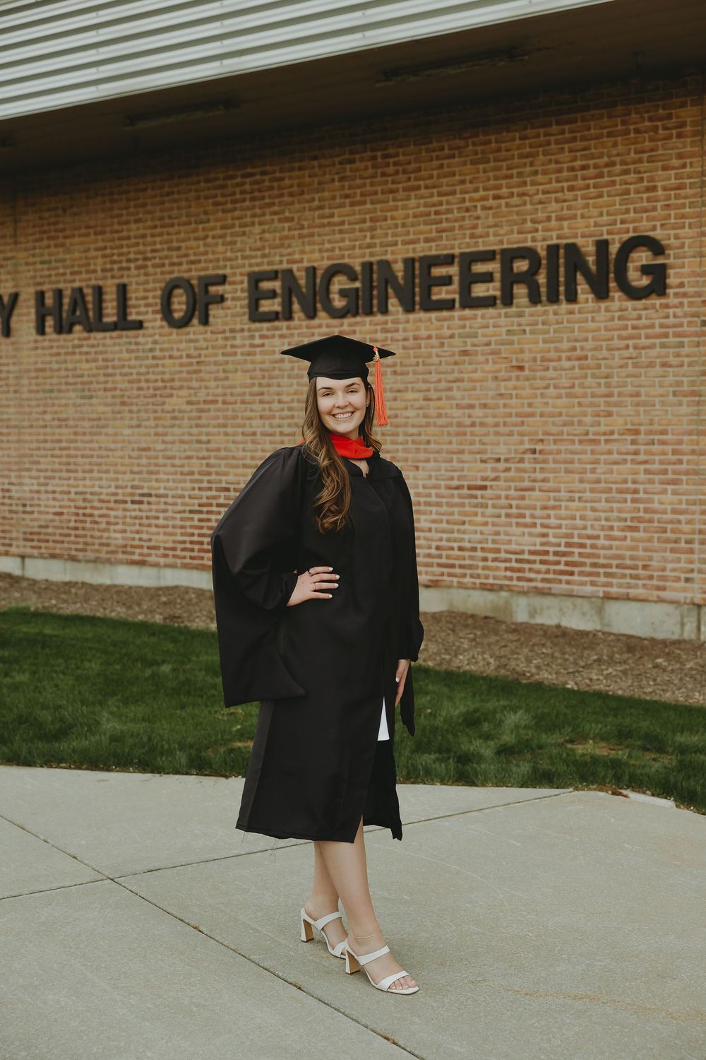 Woman in black graduation gown and cap smiles proudly in front of Hall of Engineering building.