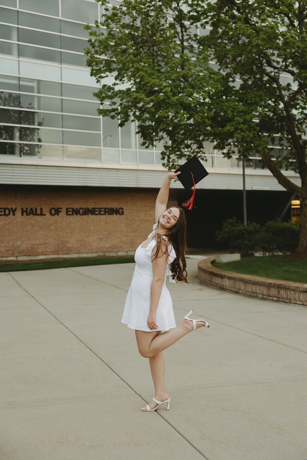 Young woman in white dress celebrates graduation by tossing cap outside Kennedy Hall of Engineering building.