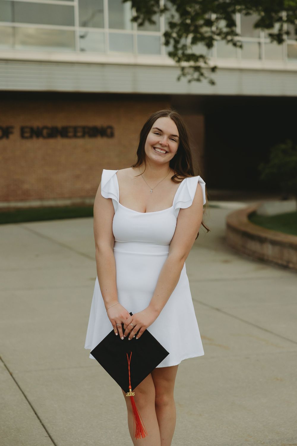 Smiling graduate in white dress holds black cap with red tassel outside College of Engineering building.