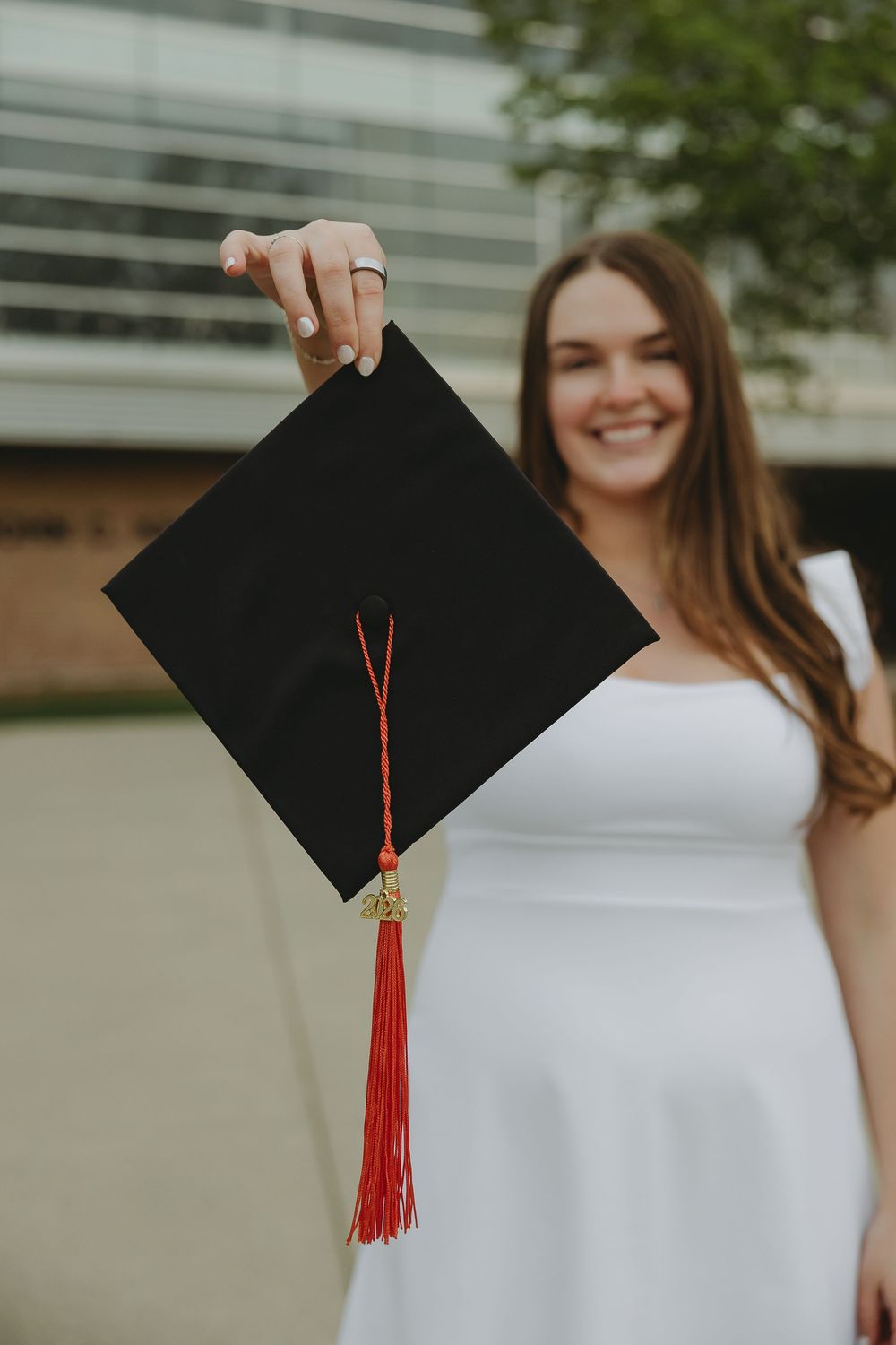 Smiling graduate in white dress holding black mortarboard cap with red tassel outdoors.