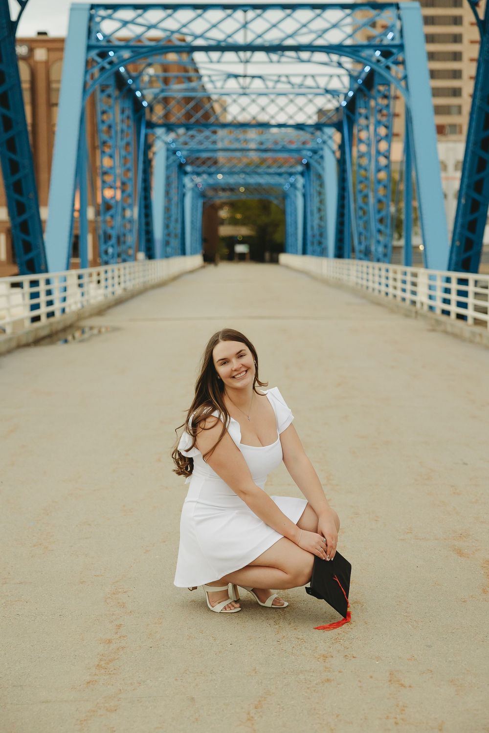 Young woman in white dress crouching on blue bridge holding graduation cap, smiling at camera.