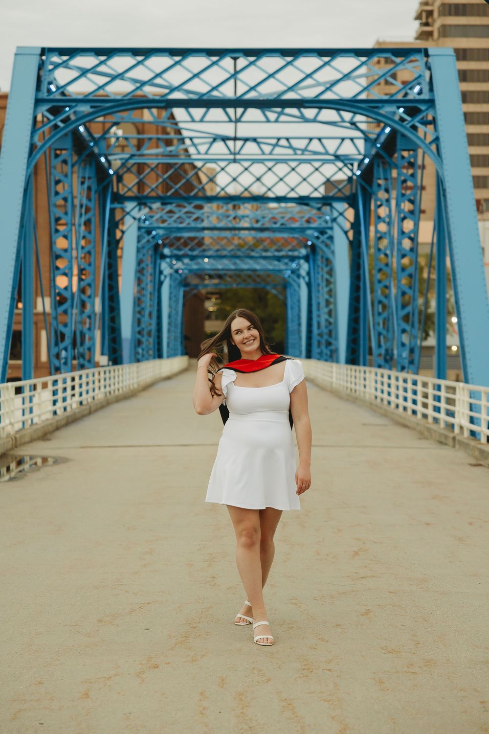 Woman in white dress and red scarf walks on blue steel bridge during graduation photo session.