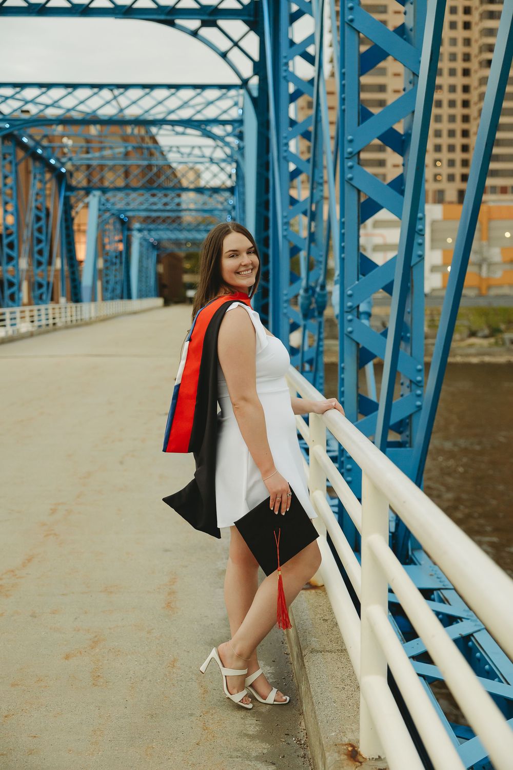 Smiling graduate in white dress holding cap on blue bridge, wearing black gown and red stole, celebrating graduation day.