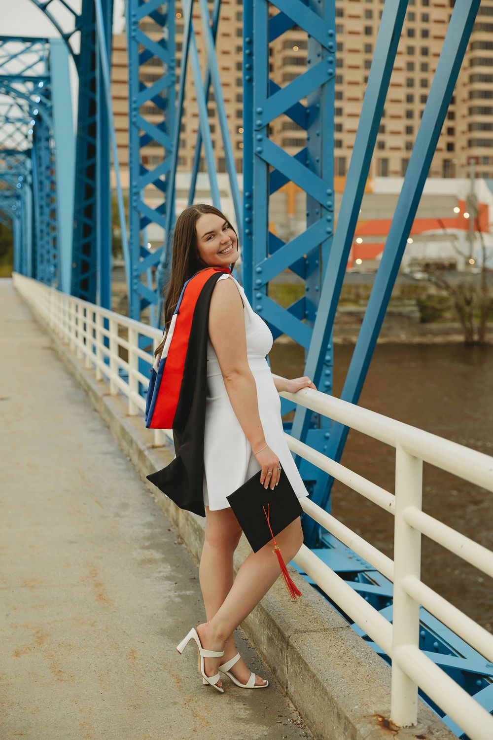 Smiling graduate in white dress holding cap and gown on blue bridge, celebrating graduation day outdoors.
