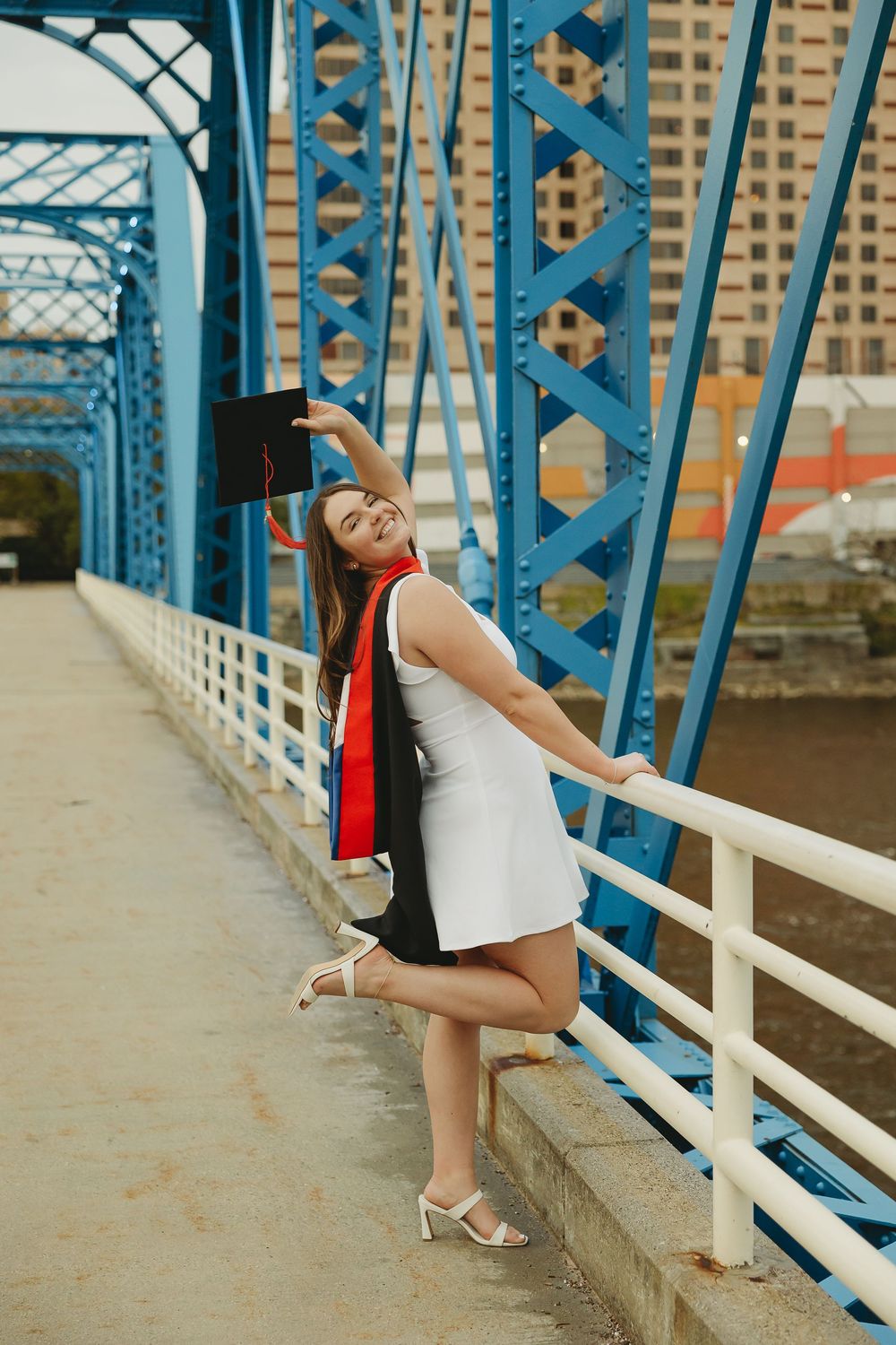 Young woman in white dress celebrating graduation on a blue steel bridge, holding cap while posing joyfully.