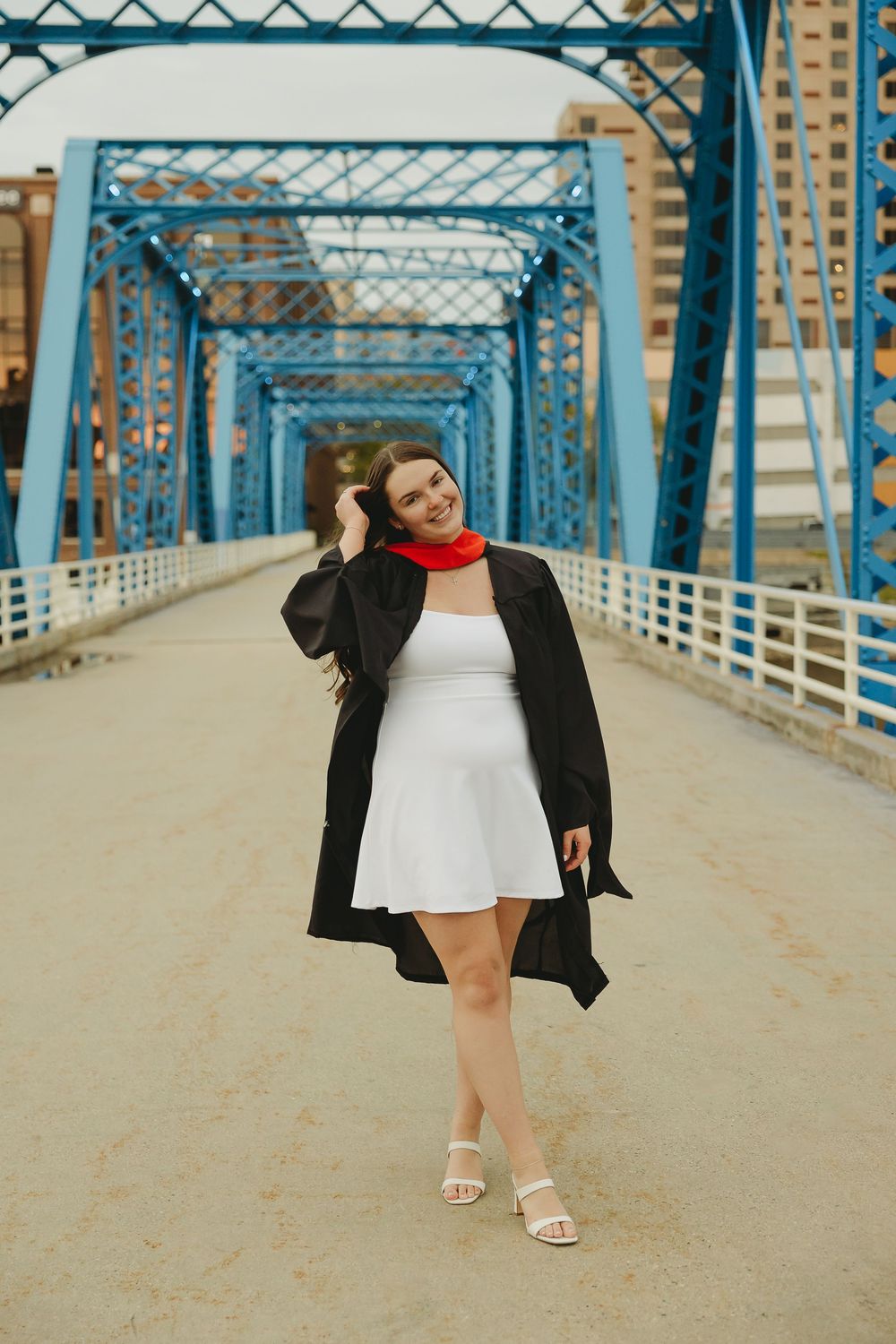 A young woman in a white dress, black graduation gown, and red scarf poses on a blue steel bridge.