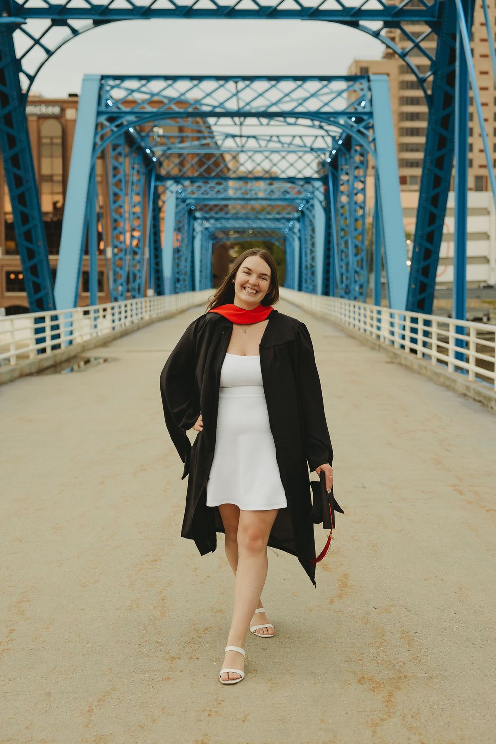 Smiling graduate in black gown and red scarf walks on blue bridge holding diploma cap.