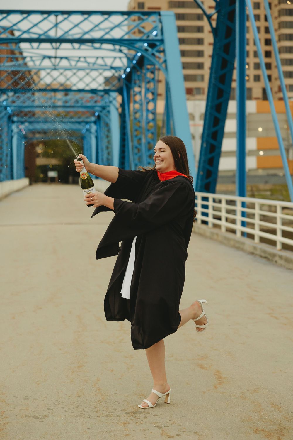 Graduating woman in black gown pops champagne on a blue bridge, celebrating her academic achievement.