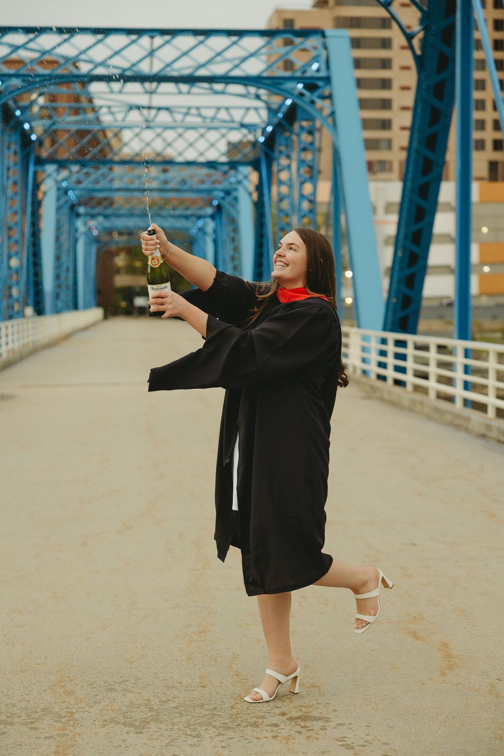 Graduate in black cap and gown celebrates on blue bridge, popping champagne while wearing white heels.