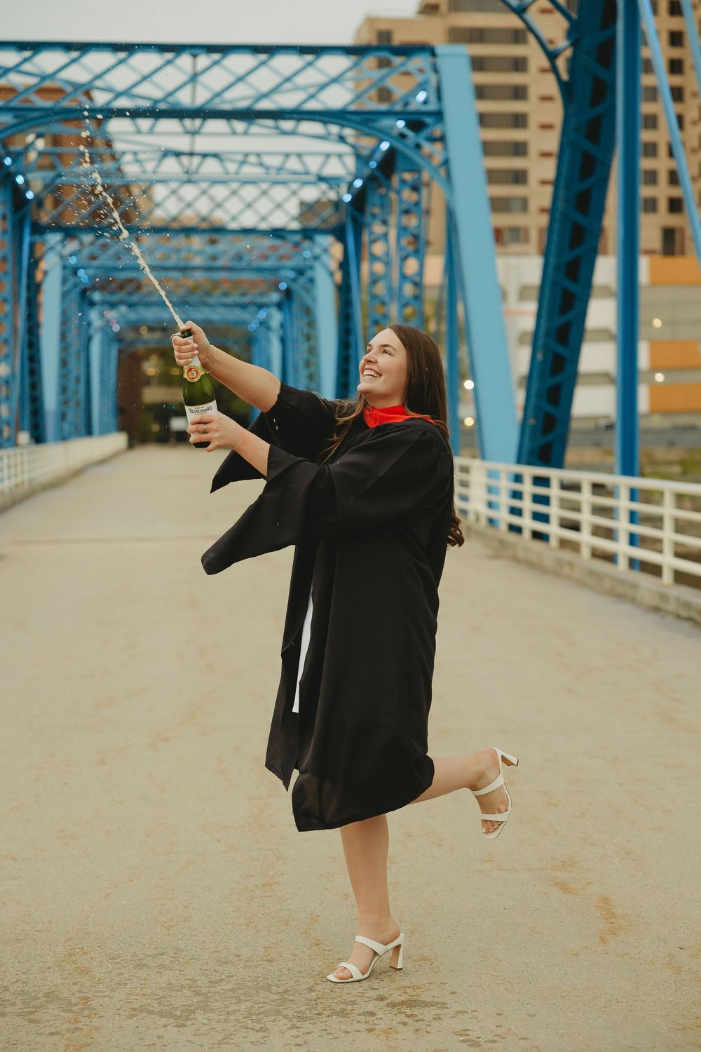 A joyful graduate in black gown celebrates on a blue bridge, holding sparkling champagne and a cup.