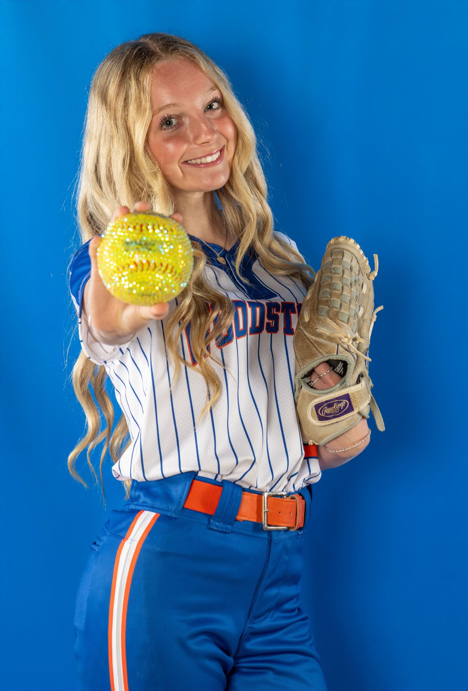 Young female softball player in blue pinstripe uniform holding yellow ball and Rawlings glove against blue background.