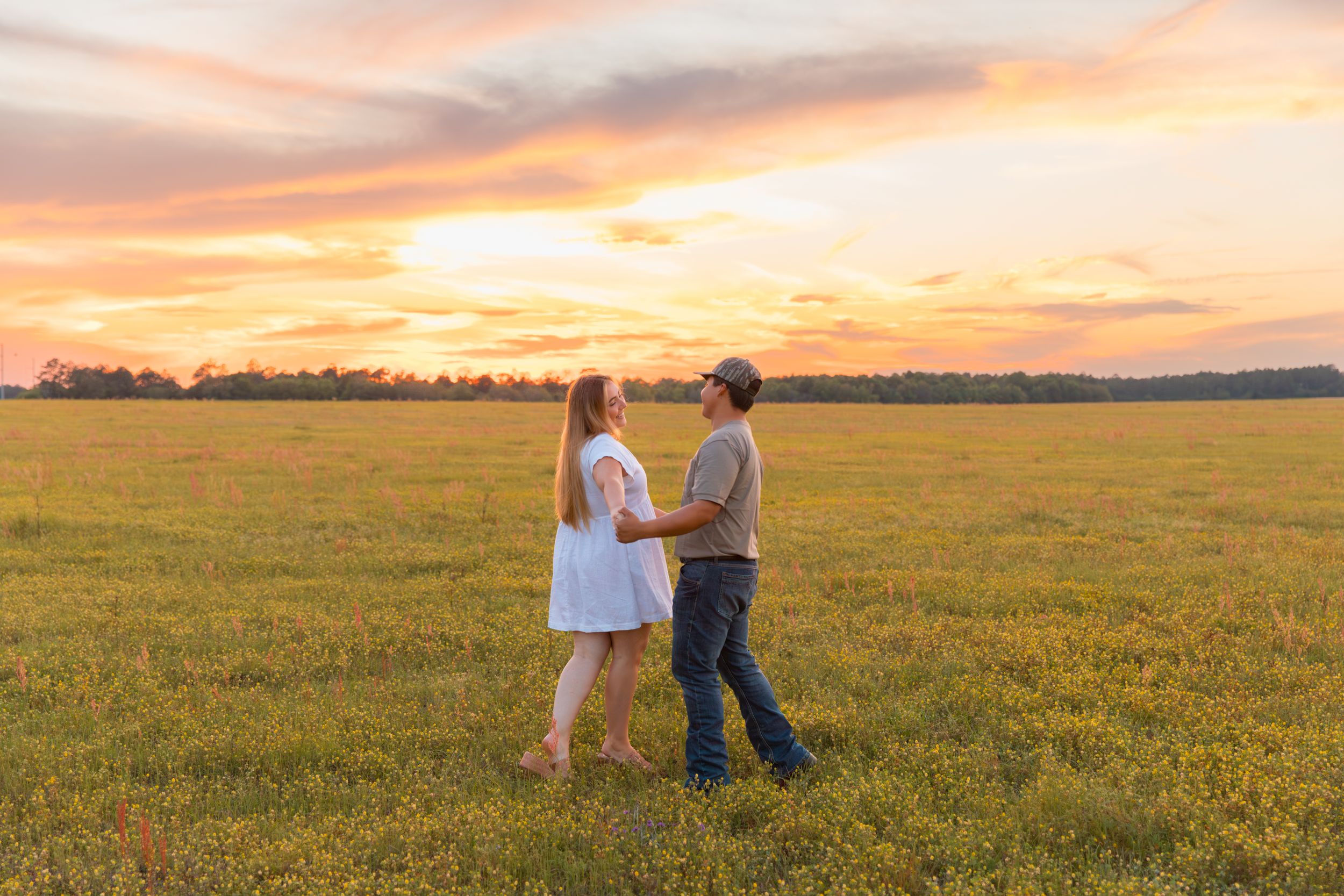 Couple holding hands in a green field at sunset with a vibrant orange and pink sky backdrop.